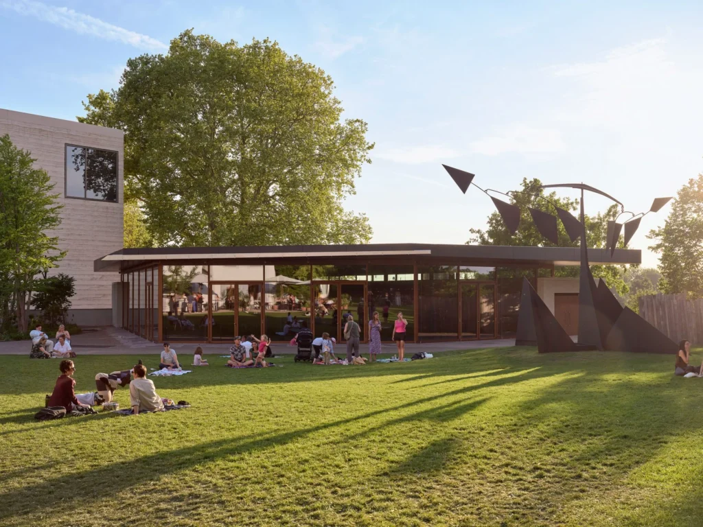 People sitting on the grass in the park with the glazed Ammann Pavilion beyond