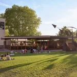 People sitting on the grass in the park with the glazed Ammann Pavilion beyond