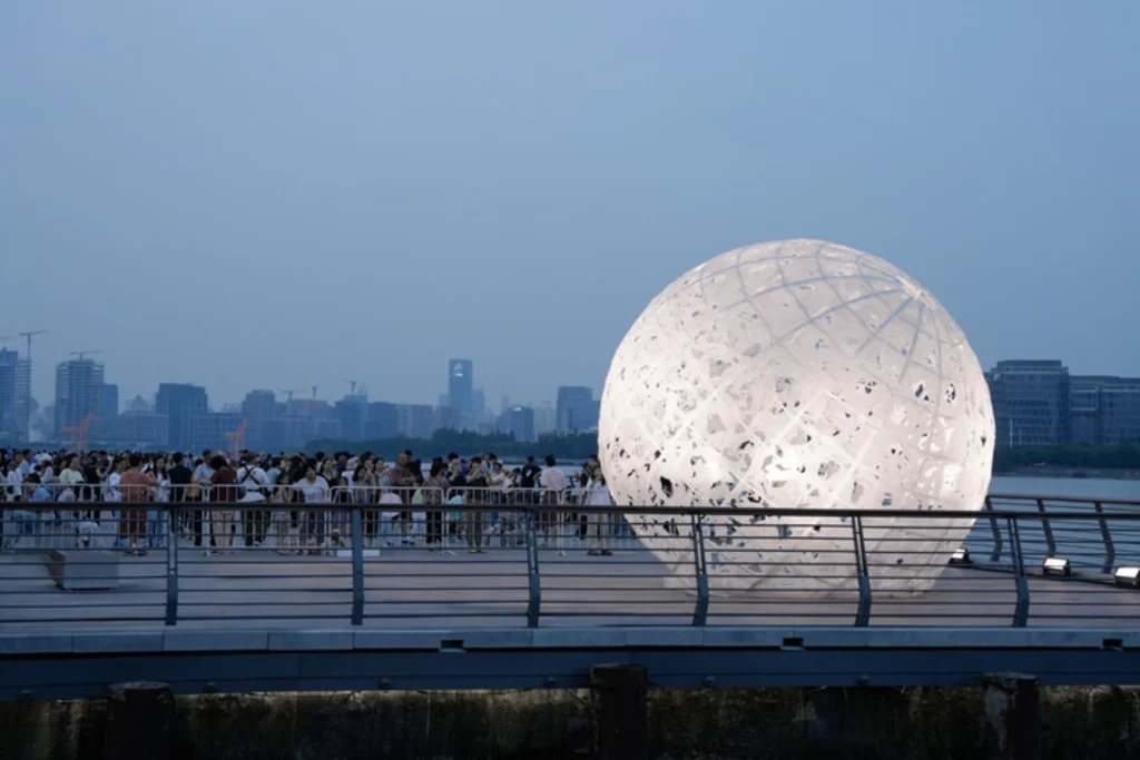 The Full Moon: Glowing spherical installation on a waterfront pier with crowds gathered behind a railing at dusk