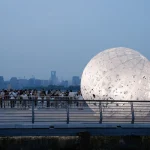 Glowing spherical installation on a waterfront pier with crowds gathered behind a railing at dusk