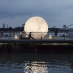 The sphere reflected in the river with a few visitors standing nearby at dusk