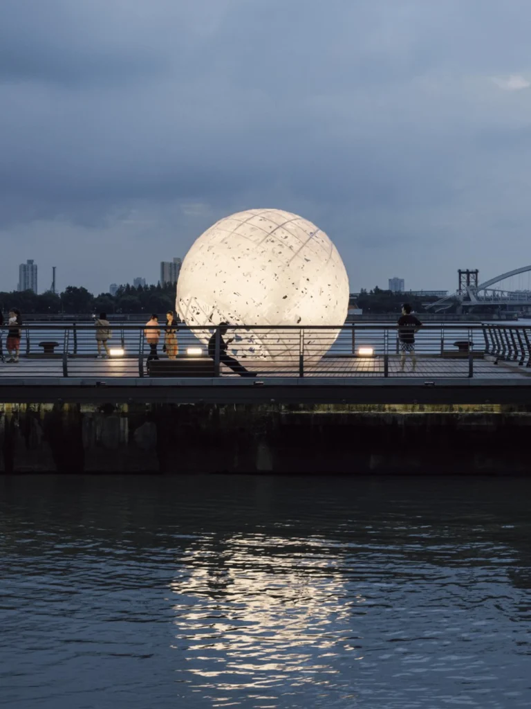 The Full Moon: The sphere reflected in the river with a few visitors standing nearby at dusk