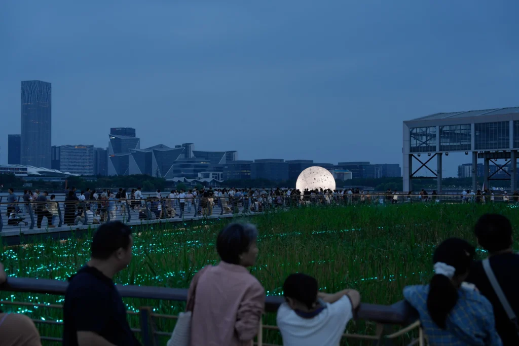 The Full Moon: Distant night view of the glowing sphere across reeds and a boardwalk filled with people
