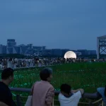 Distant night view of the glowing sphere across reeds and a boardwalk filled with people