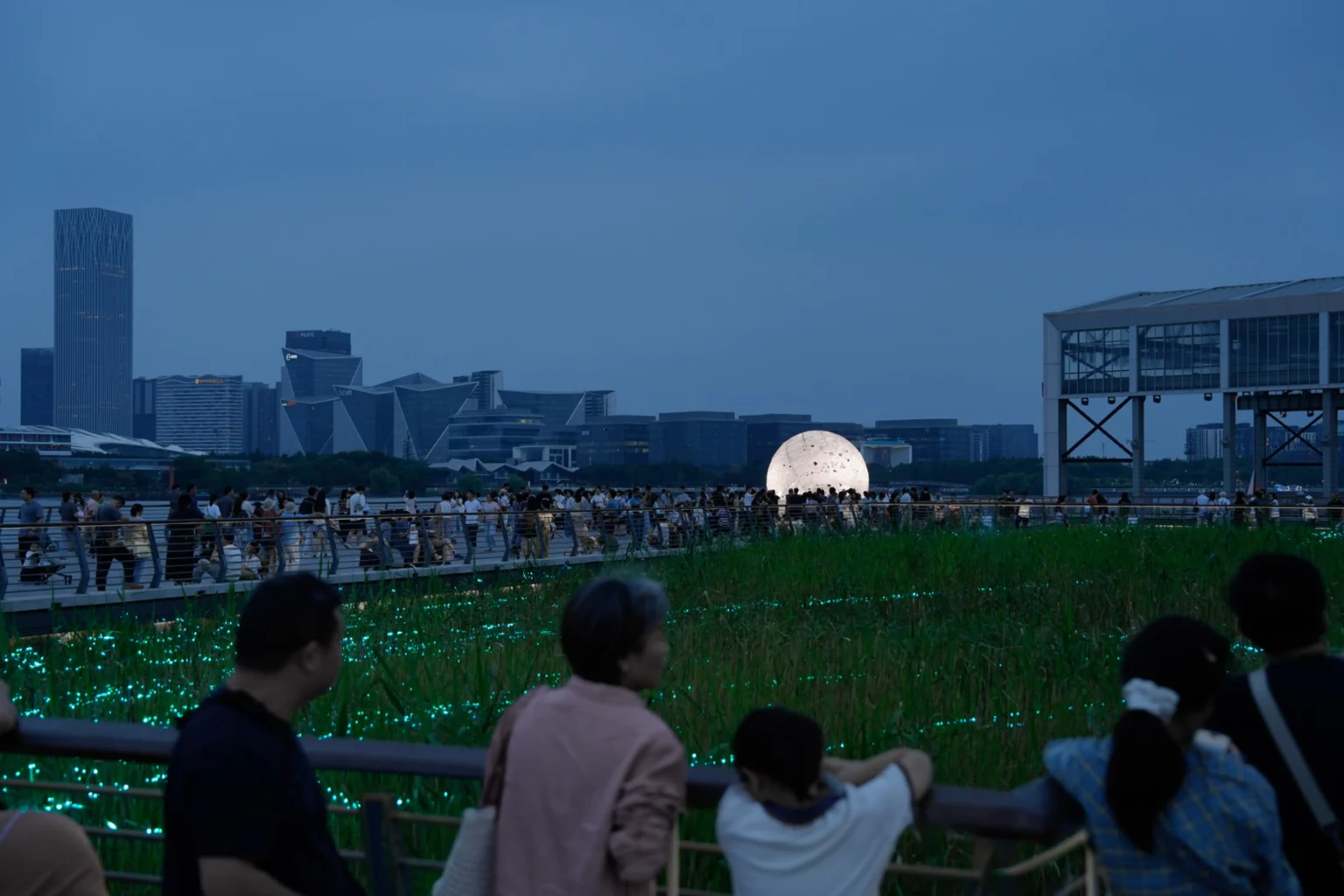 Distant night view of the glowing sphere across reeds and a boardwalk filled with people