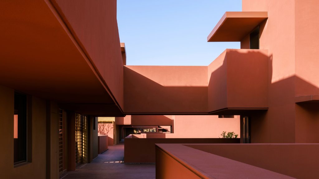 Narrow terracotta-coloured walkway framed by overhanging volumes under clear blue sky