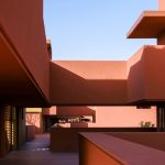 Narrow terracotta-coloured walkway framed by overhanging volumes under clear blue sky