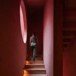 View up a coral-red staircase toward a circular window with a person standing at the top