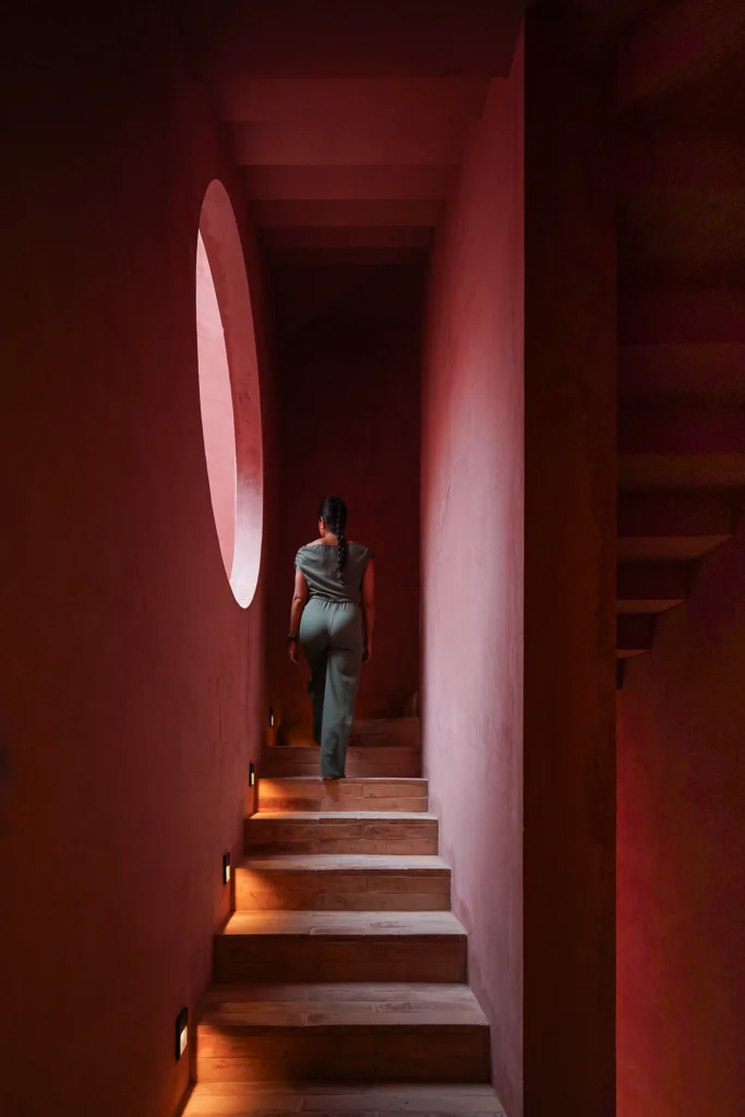 View up a coral-red staircase toward a circular window with a person standing at the top