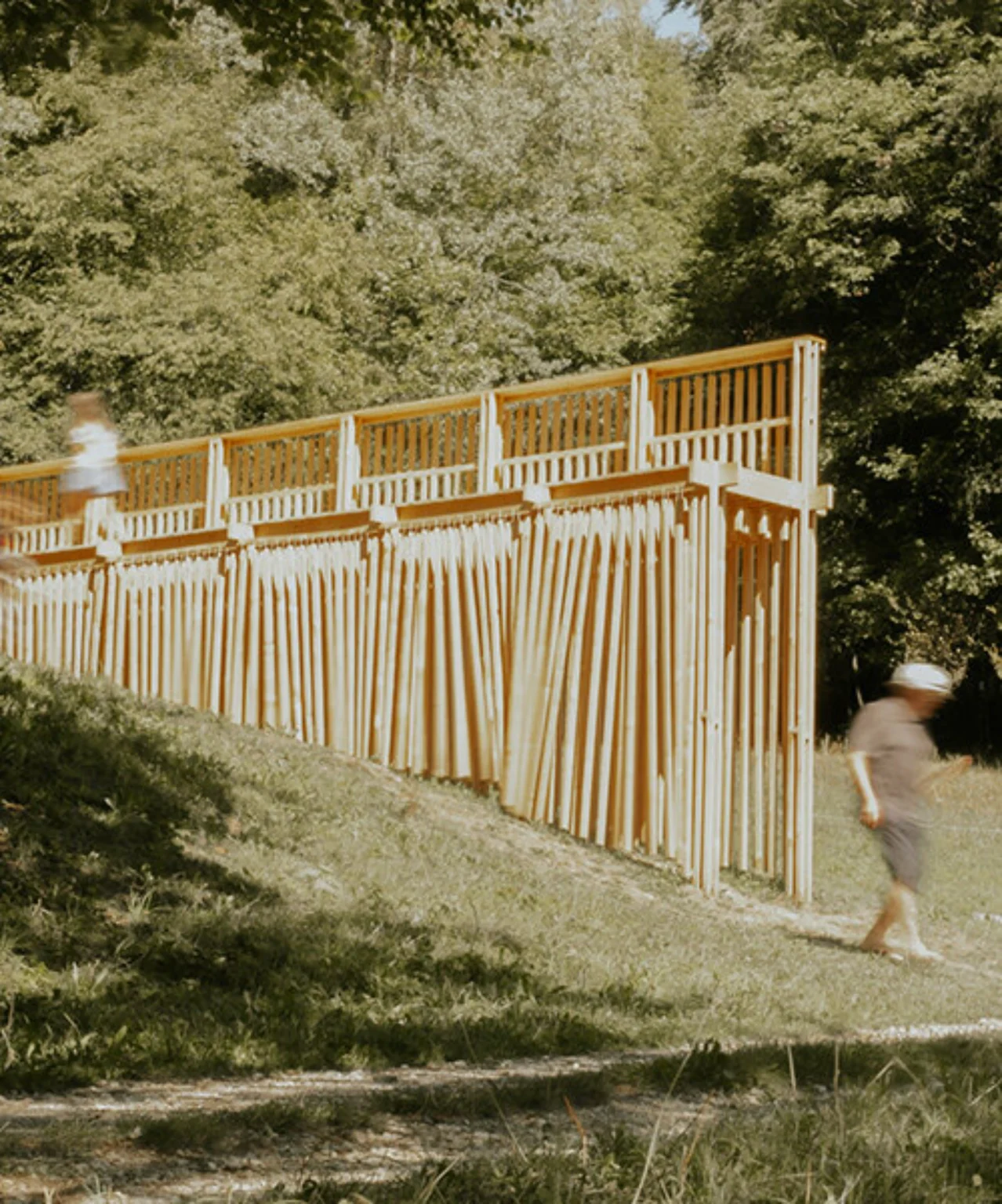 Person walking beside the long timber pavilion with vertical wooden elements on a grassy Alpine slope