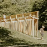 Person walking beside the long timber pavilion with vertical wooden elements on a grassy Alpine slope