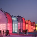 Row of illuminated arched fabric pavilions in pink, purple, blue, red, orange and yellow glowing at dusk