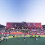 Large red fabric-clad stadium structure with crowds gathering on colorful ground surfaces at twilight