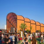 Long orange arched pavilion under clear blue sky with crowds moving along the entrance area