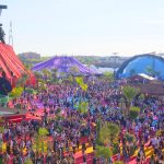 Wide aerial view of dense crowds on rainbow pathways with purple and blue fabric structures beyond