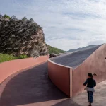 Young child running along a curved reddish path beside a large woven organic structure