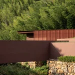 Reddish architectural volumes set against dense bamboo forest and a stone retaining wall
