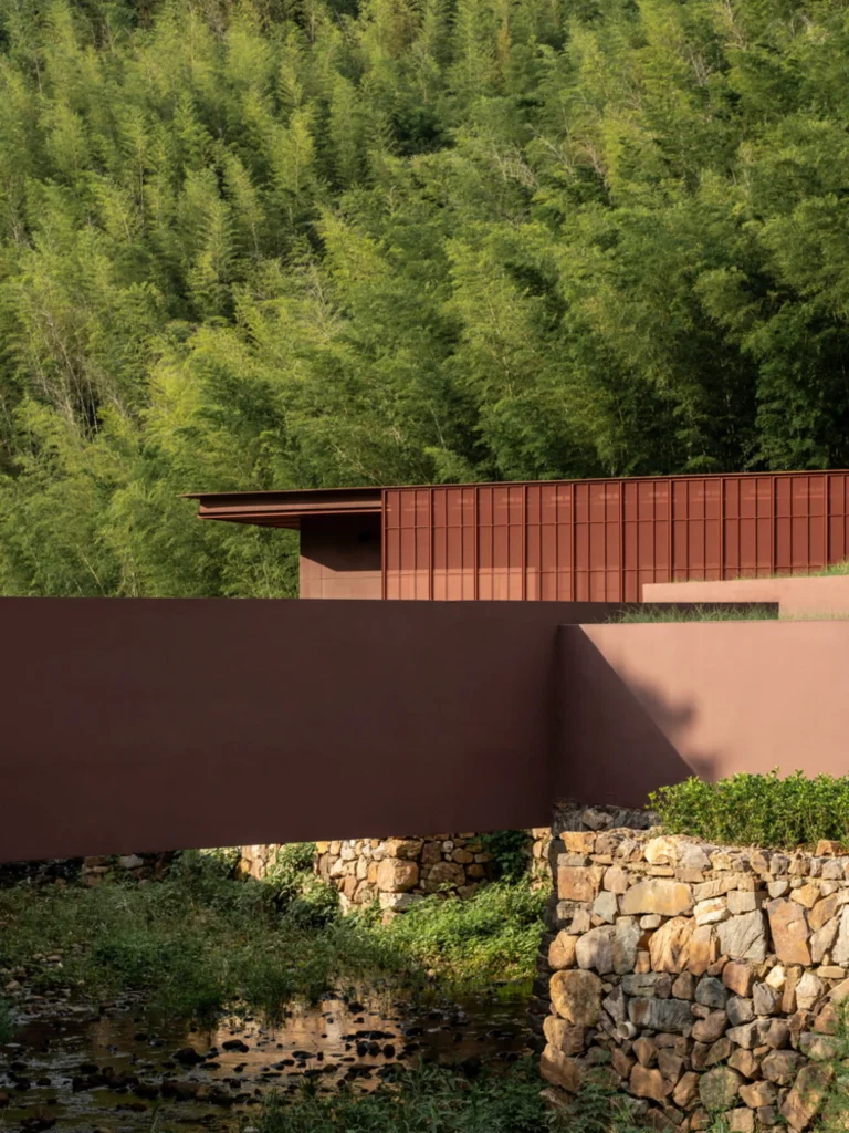 Earth Valley Theater: Reddish architectural volumes set against dense bamboo forest and a stone retaining wall