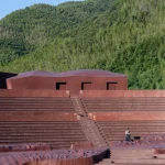 Reddish architectural volumes set against dense bamboo forest and a stone retaining wall