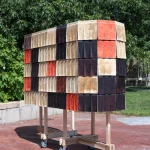 Outdoor view of a tall, rectangular wooden-framed structure on casters, clad in overlapping fluted tiles of beige, black, red, and brown wool biocomposite, standing on a red-brick plaza with green trees and a building in the background.