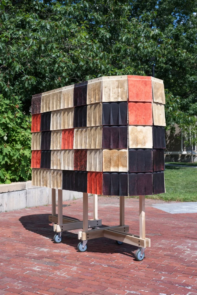 Outdoor view of a tall, rectangular wooden-framed structure on casters, clad in overlapping fluted tiles of beige, black, red, and brown wool biocomposite, standing on a red-brick plaza with green trees and a building in the background.
