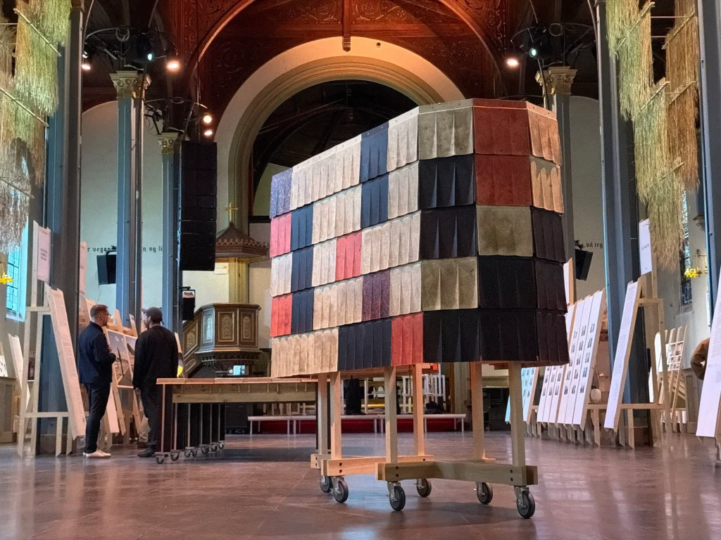 Harvard: Interior view inside a historic church with high vaulted ceiling and arches, showing the same tall rectangular structure clad in multicolored fluted wool tiles on a wheeled wooden base, with visitors and exhibition panels nearby.