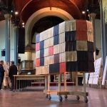 Interior view inside a historic church with high vaulted ceiling and arches, showing the same tall rectangular structure clad in multicolored fluted wool tiles on a wheeled wooden base, with visitors and exhibition panels nearby.