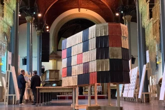 Interior view inside a historic church with high vaulted ceiling and arches, showing the same tall rectangular structure clad in multicolored fluted wool tiles on a wheeled wooden base, with visitors and exhibition panels nearby.