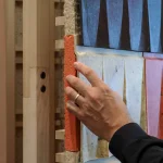 Close-up of a hand touching the layered edge of the installation, revealing the thick orange outer wool-tile face, denser inner layers, and soft fibrous insulation against a light timber frame.