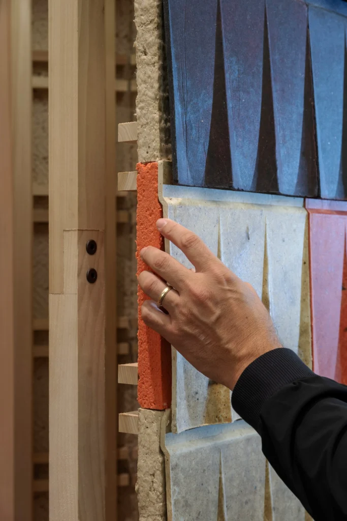 Close-up of a hand touching the layered edge of the installation, revealing the thick orange outer wool-tile face, denser inner layers, and soft fibrous insulation against a light timber frame.