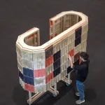 Overhead view of a large C-shaped wooden structure on casters, fully clad in a checkerboard of beige, black, red, and brown fluted wool tiles, with a person in a black shirt standing beside it and touching the surface in a dark exhibition hall.