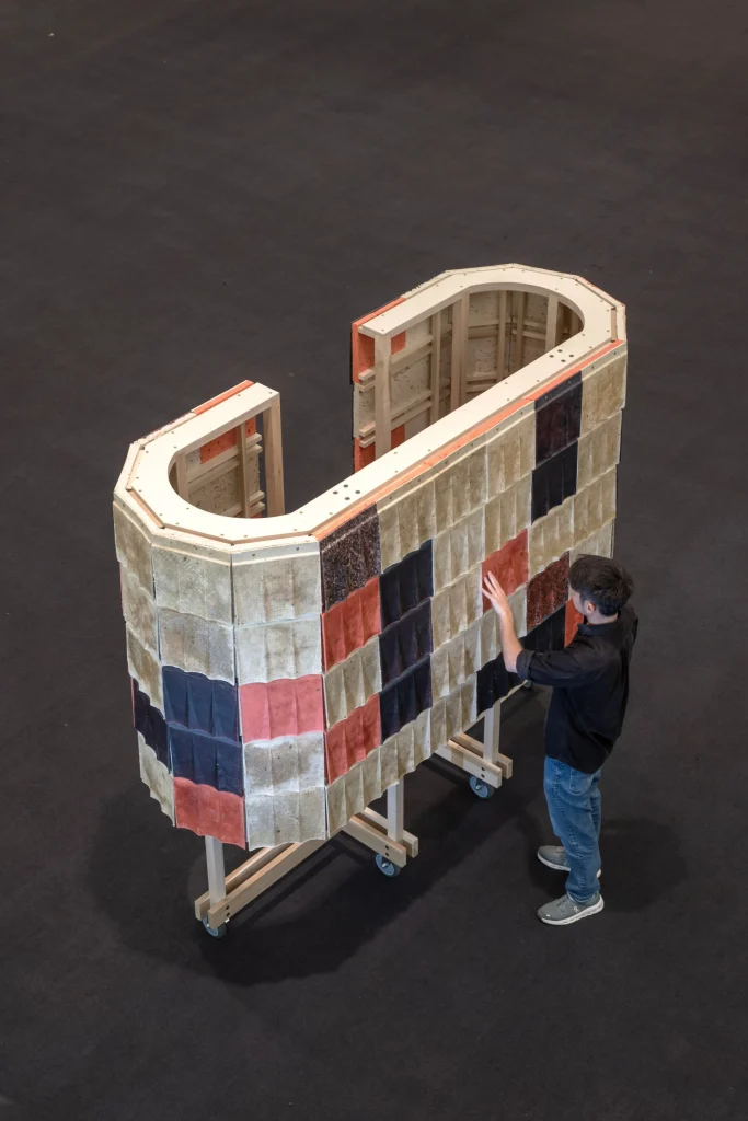 Overhead view of a large C-shaped wooden structure on casters, fully clad in a checkerboard of beige, black, red, and brown fluted wool tiles, with a person in a black shirt standing beside it and touching the surface in a dark exhibition hall.