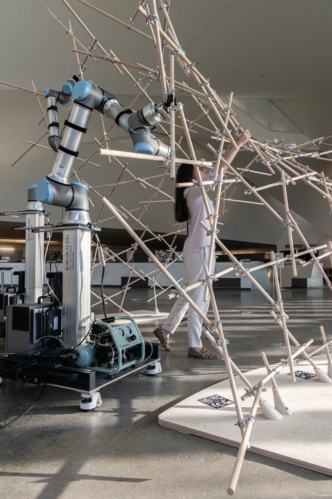 Futuristic illustration of a human designer and collaborative robots working together on a construction site. A glowing digital blueprint floats between them, transforming into physical robot-ready assembly instructions as robotic arms precisely place structural components under AI guidance.