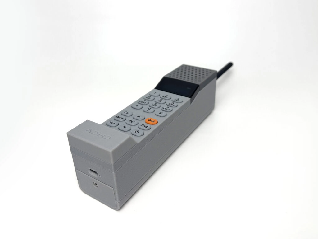 Side view of the grey 3D-printed ADHD ONE handset showing the keypad, orange Send button and charging port