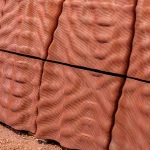 Close-up of reddish-brown 3D-printed wall panels with a ribbed, undulating surface texture resting on red soil