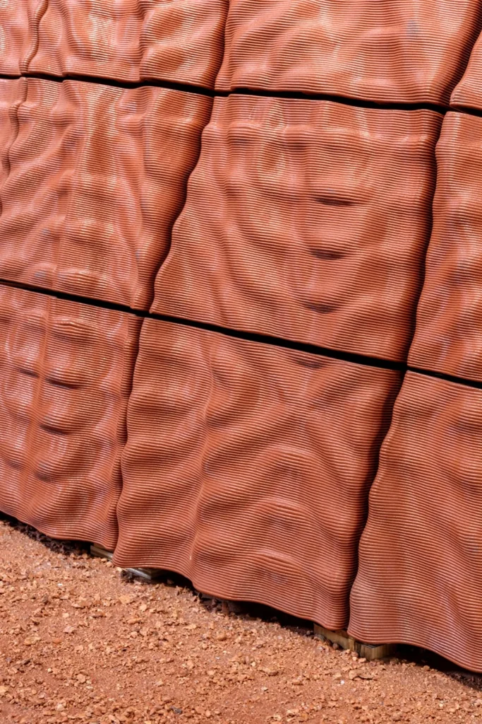 Close-up of reddish-brown 3D-printed wall panels with a ribbed, undulating surface texture resting on red soil