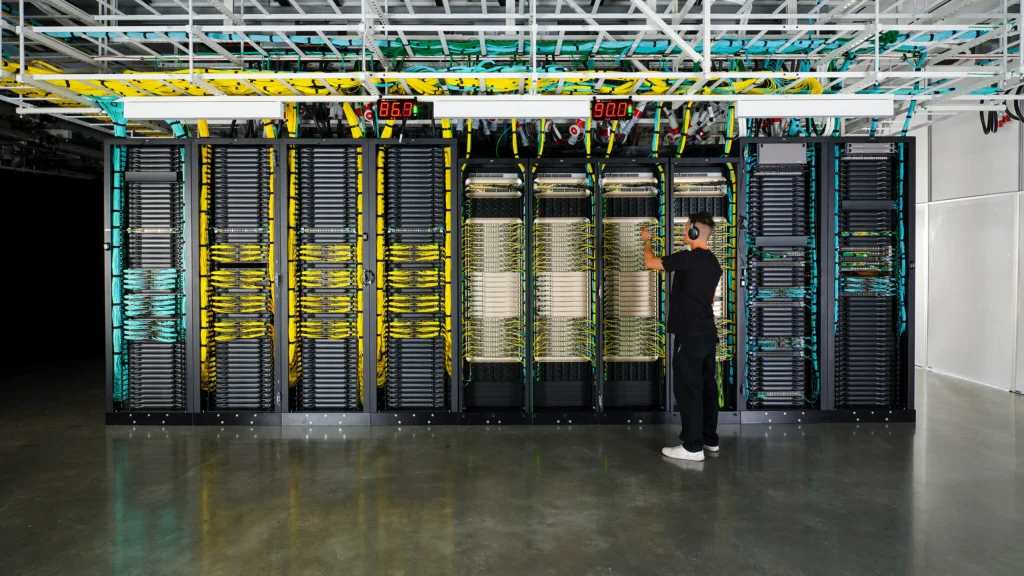 Technician in a black T-shirt working on open server racks filled with yellow and teal network cables inside a modern data centre