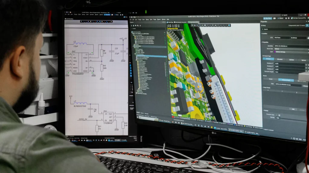 Close-up of a designer working on dual screens showing circuit schematics on the left and a colourful 3D PCB assembly model on the right