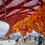 A vibrant red lattice canopy roof covering a pedestrian walkway, featuring suspended layers of orange and yellow fabric flags above visitors.