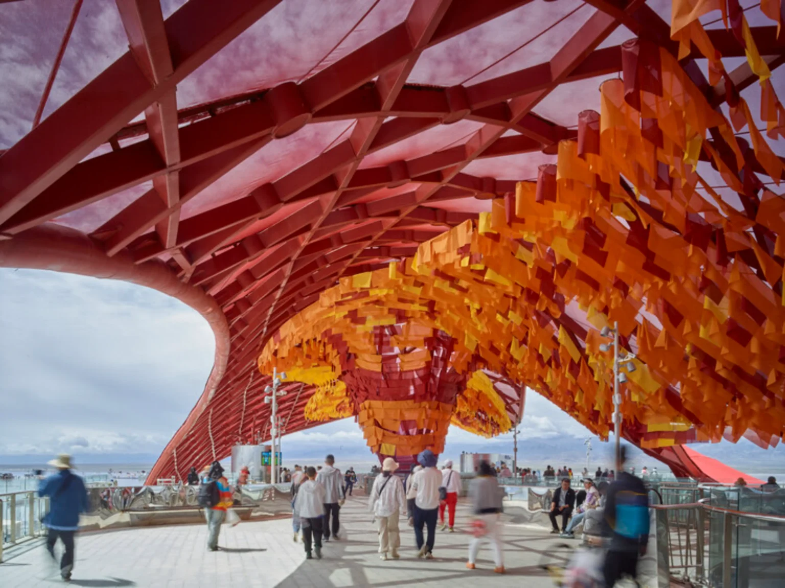 A vibrant red lattice canopy roof covering a pedestrian walkway, featuring suspended layers of orange and yellow fabric flags above visitors.