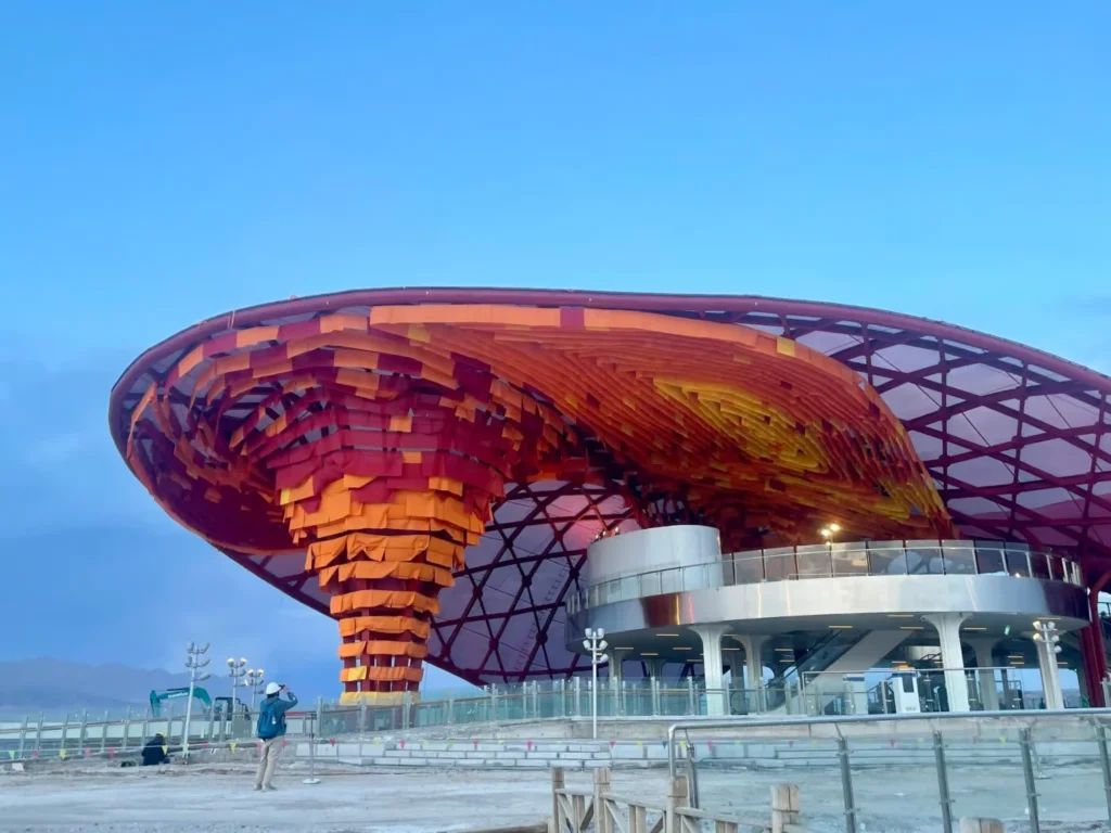 Exterior view of an architectural pavilion at dusk showing a central inverted funnel structure wrapped in layers of orange, yellow, and red fabric.