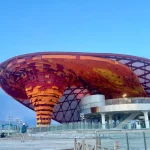Exterior view of an architectural pavilion at dusk showing a central inverted funnel structure wrapped in layers of orange, yellow, and red fabric.
