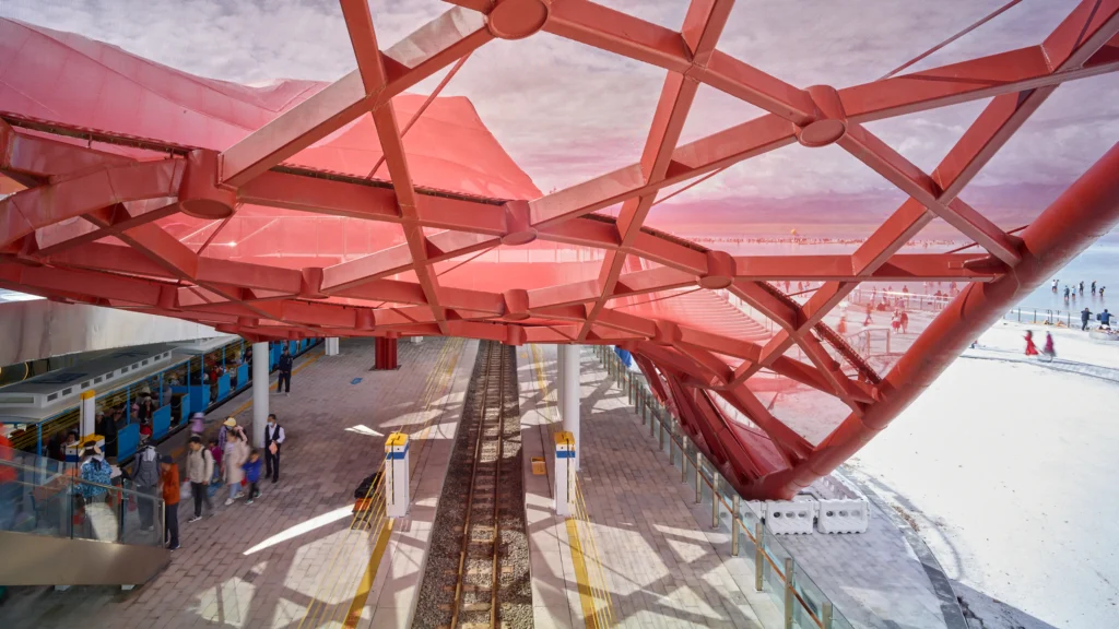 Eye-level view beneath a red structural frame over train tracks and a platform filled with passengers next to a white salt flat landscape.