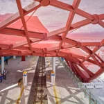 Eye-level view beneath a red structural frame over train tracks and a platform filled with passengers next to a white salt flat landscape.