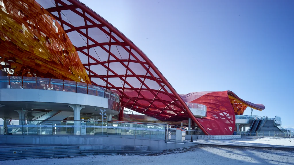 Side elevation of a dynamic red lattice roof structure sweeping downward toward the ground, set against a bright blue sky.