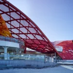 Side elevation of a dynamic red lattice roof structure sweeping downward toward the ground, set against a bright blue sky.