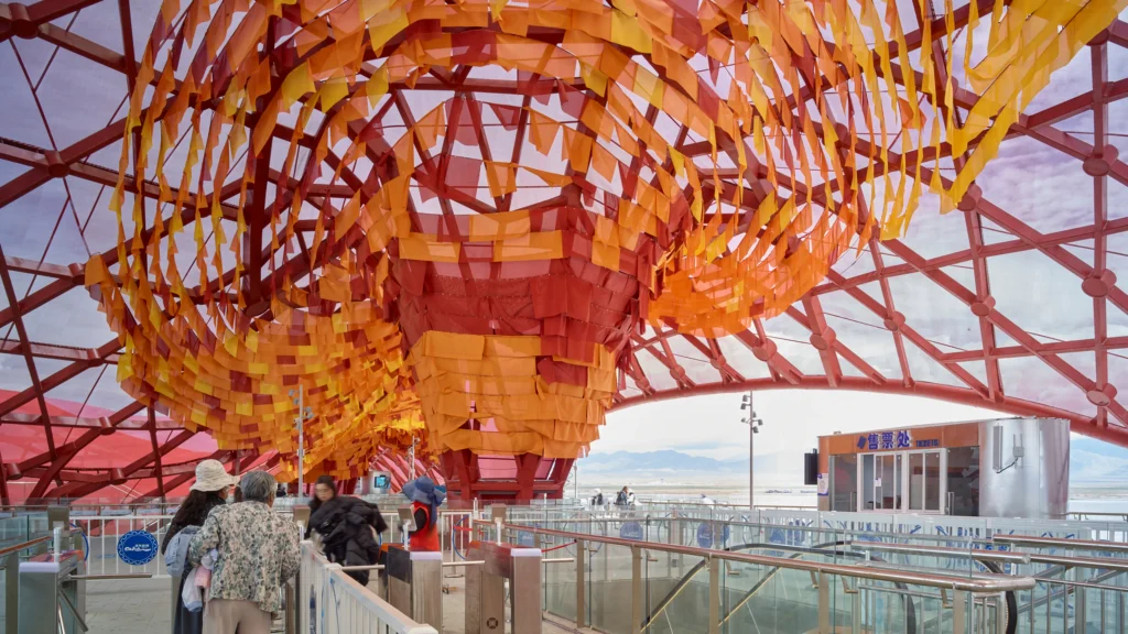 Interior view of turnstiles and ticket booth underneath a massive, funnel-shaped ceiling installation made of orange and yellow fabric strips.