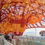 Interior view of turnstiles and ticket booth underneath a massive, funnel-shaped ceiling installation made of orange and yellow fabric strips.