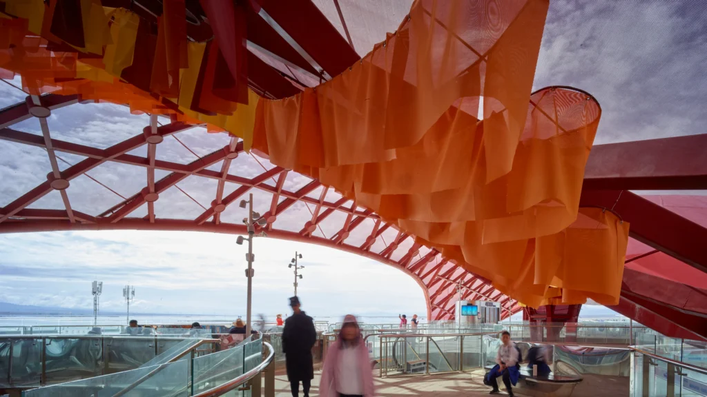Pedestrians walking along a curved glass-railed walkway beneath hanging orange fabric sheets and a red lattice canopy.
