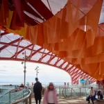 Pedestrians walking along a curved glass-railed walkway beneath hanging orange fabric sheets and a red lattice canopy.